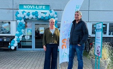 Foto: Mit Luftballons geschmückter Eingangsbereich des Kaufhauses NOVI-LIFE. Davor zwei Personen mit einem Beach-flag zum 20-Jährigen. 