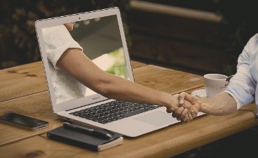Digital verbunden - gemeinsam stark! Aus einem Laptopbildschirm ragt ein Arm heraus, dessen Hand eine andere Hand außerhalb des Laptops hält.