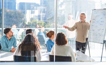 Fünf Personen sitzen um einen Tisch und folgen den Ausführungen eines Dozenten am Whiteboard.