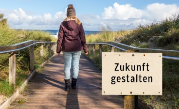 Beispielbild: Eine Frau läuft warm angezogen auf einem Holzsteg dem Meer entgegen. Auf einem Schild steht: Zukunft gestalten. 