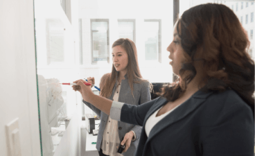 Beispielfoto: Zwei Menschen diskutieren und schreiben gemeinsam an ein Whiteboard im hellen Büro.