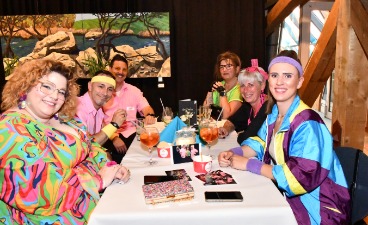 Foto von der 80er Jahre Party: An einem Tisch sitzen sich fröhliche, im 80er Jahre Stil angezogene, Menschen gegenüber. 