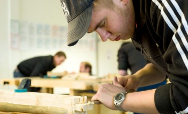 Beispielfoto: Im Vordergrund arbeitet ein junger Mann an der Werkbank, im Hintergrund sprechen zwei Männer miteinander.
