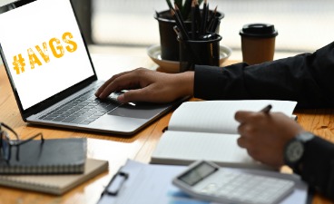 Beispielbild: Person arbeitet konzentriert am Laptop mit #AVGS auf dem Bildschirm, schreibt Notizen und nutzt Bürobedarf auf einem Schreibtisch.